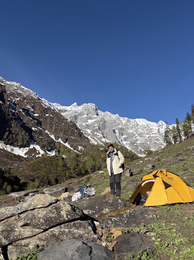 Campsite with a yellow tent below snow-covered peaks at Beas Kund, Himachal